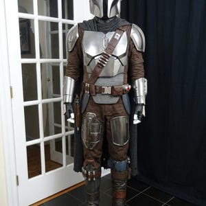 May include: A full-body costume resembling a character from a science fiction series. The costume features a silver helmet, chest armor, and brown and gray pants. A brown leather belt and shoulder strap are also visible. The costume includes a cape and gloves.