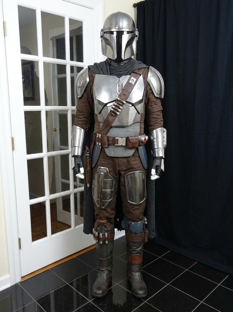 May include: A full-body costume resembling a character from a science fiction series. The costume features a silver helmet, chest armor, and brown and gray pants. A brown leather belt and shoulder strap are also visible. The costume includes a cape and gloves.