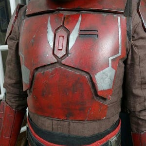 Puede incluir: Un peto de armadura mandaloriana rojo y plateado desgastado con un traje interior marr&oacute;n. El peto presenta un dise&ntilde;o rectangular con detalles plateados y un acabado desgastado, lo que sugiere desgaste de batalla. Parte de un disfraz.