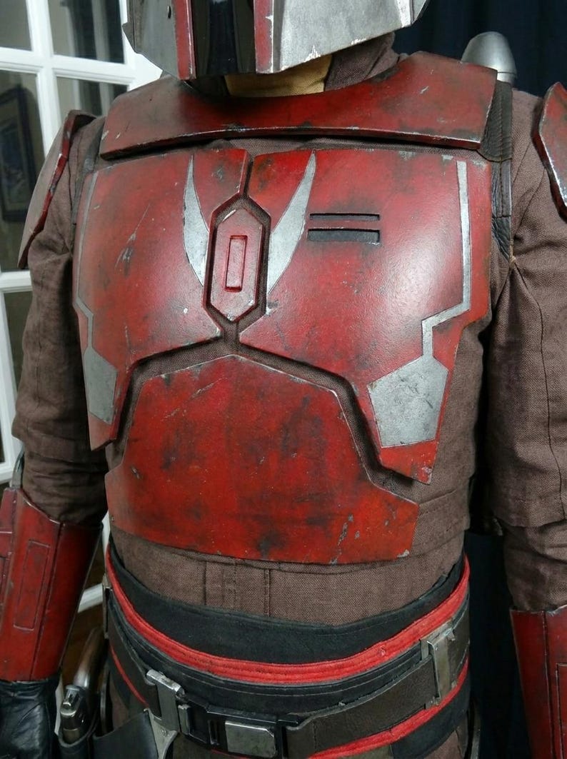 Puede incluir: Un peto de armadura mandaloriana rojo y plateado desgastado con un traje interior marr&oacute;n. El peto presenta un dise&ntilde;o rectangular con detalles plateados y un acabado desgastado, lo que sugiere desgaste de batalla. Parte de un disfraz.