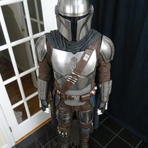 May include: A full-body costume resembling the Mandalorian from the Star Wars series. The costume features a silver helmet, chest armor, and shoulder pads. It includes a brown and gray jumpsuit, a utility belt with pouches, and boots. The costume is designed to replicate the character's iconic look.