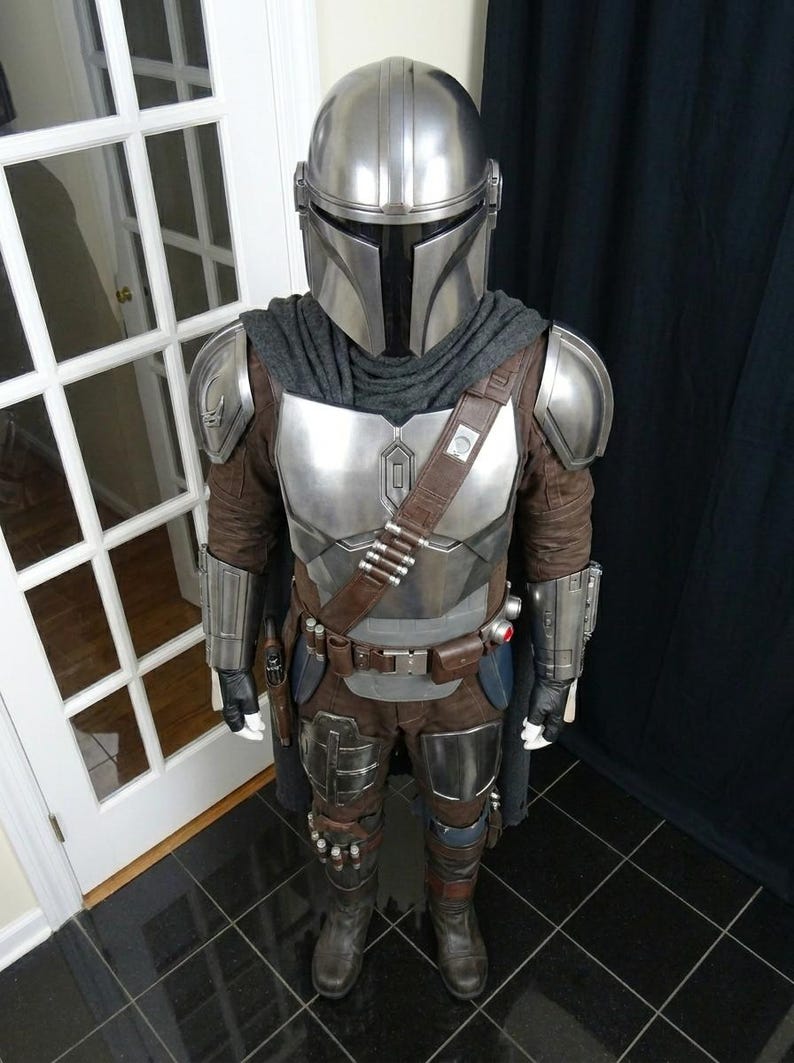 May include: A full-body costume resembling the Mandalorian from the Star Wars series. The costume features a silver helmet, chest armor, and shoulder pads. It includes a brown and gray jumpsuit, a utility belt with pouches, and boots. The costume is designed to replicate the character's iconic look.