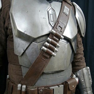 May include: A detailed costume featuring a metallic silver chest plate, shoulder armor, and arm gauntlet. A brown leather belt and shoulder strap hold ammunition and a small pouch. The costume includes a brown jacket and a gray scarf.