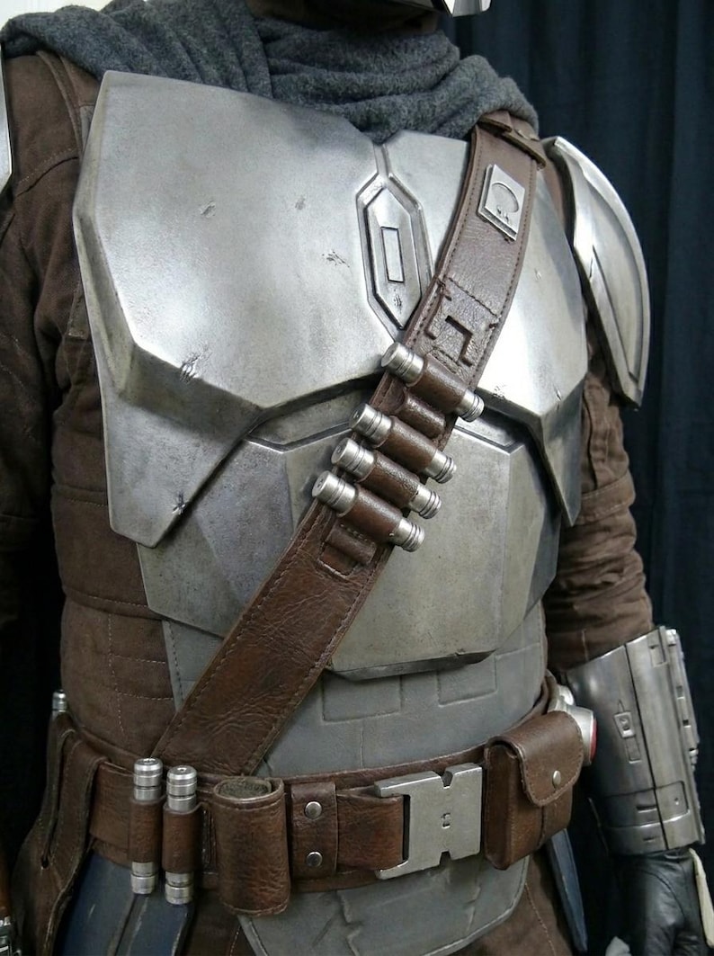 May include: A detailed costume featuring a metallic silver chest plate, shoulder armor, and arm gauntlet. A brown leather belt and shoulder strap hold ammunition and a small pouch. The costume includes a brown jacket and a gray scarf.