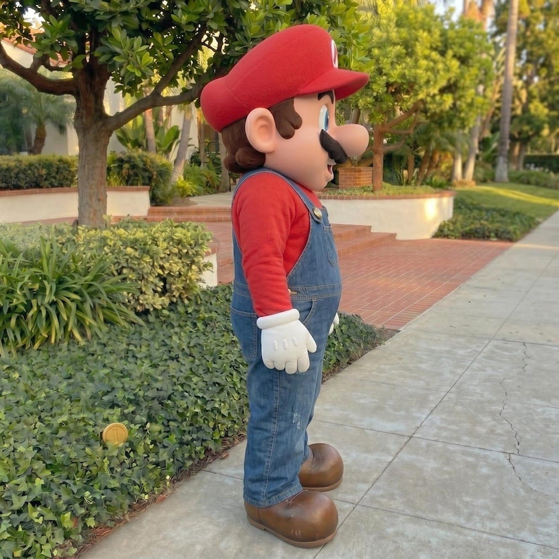Pu&ograve; includere: Un costume integrale del personaggio del videogioco Mario. Il costume presenta un cappello rosso, una maglietta rossa a maniche lunghe, una salopette di jeans blu, guanti bianchi e scarpe marroni. Il personaggio &egrave; in piedi su un marciapiede.