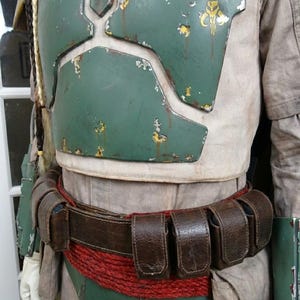 May include: A detailed costume featuring a weathered green chest plate with yellow accents, a brown leather belt with pouches, and a red rope belt. The costume has a worn appearance, suggesting use.