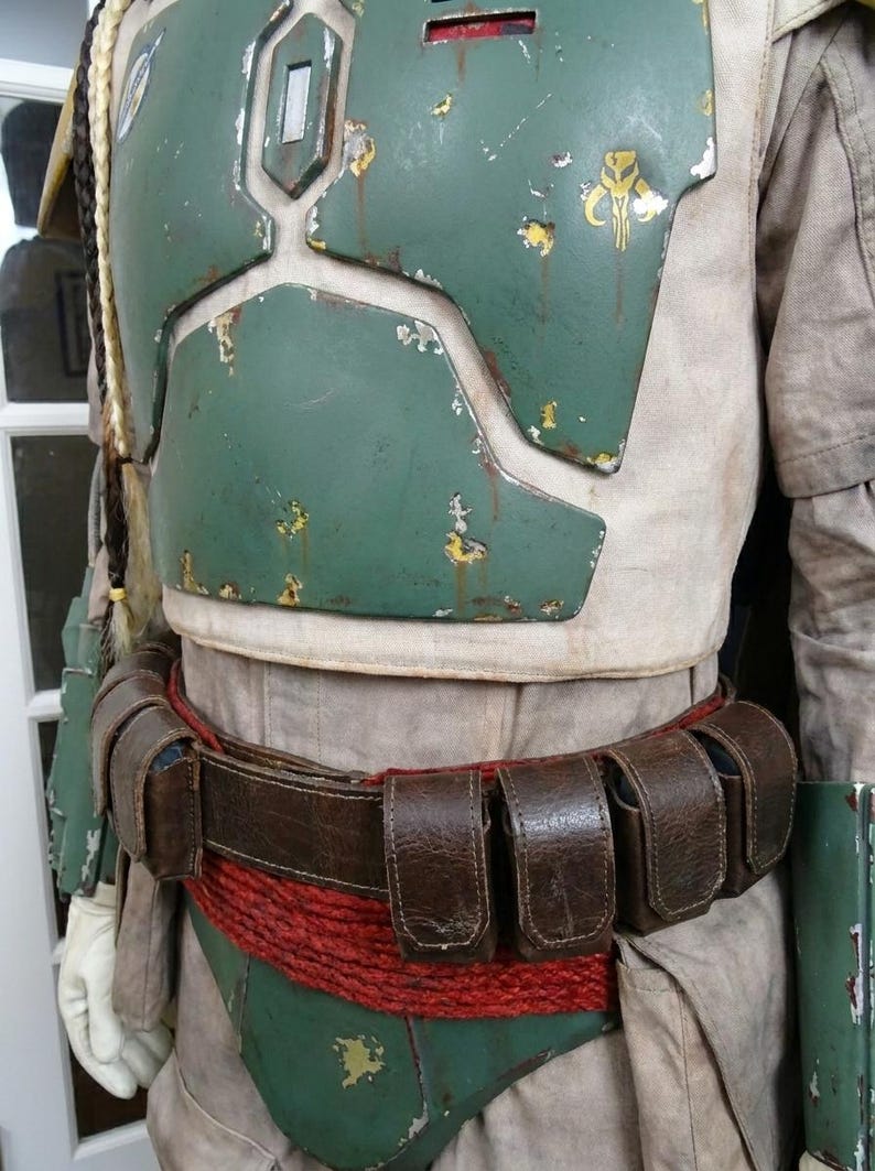 May include: A detailed costume featuring a weathered green chest plate with yellow accents, a brown leather belt with pouches, and a red rope belt. The costume has a worn appearance, suggesting use.