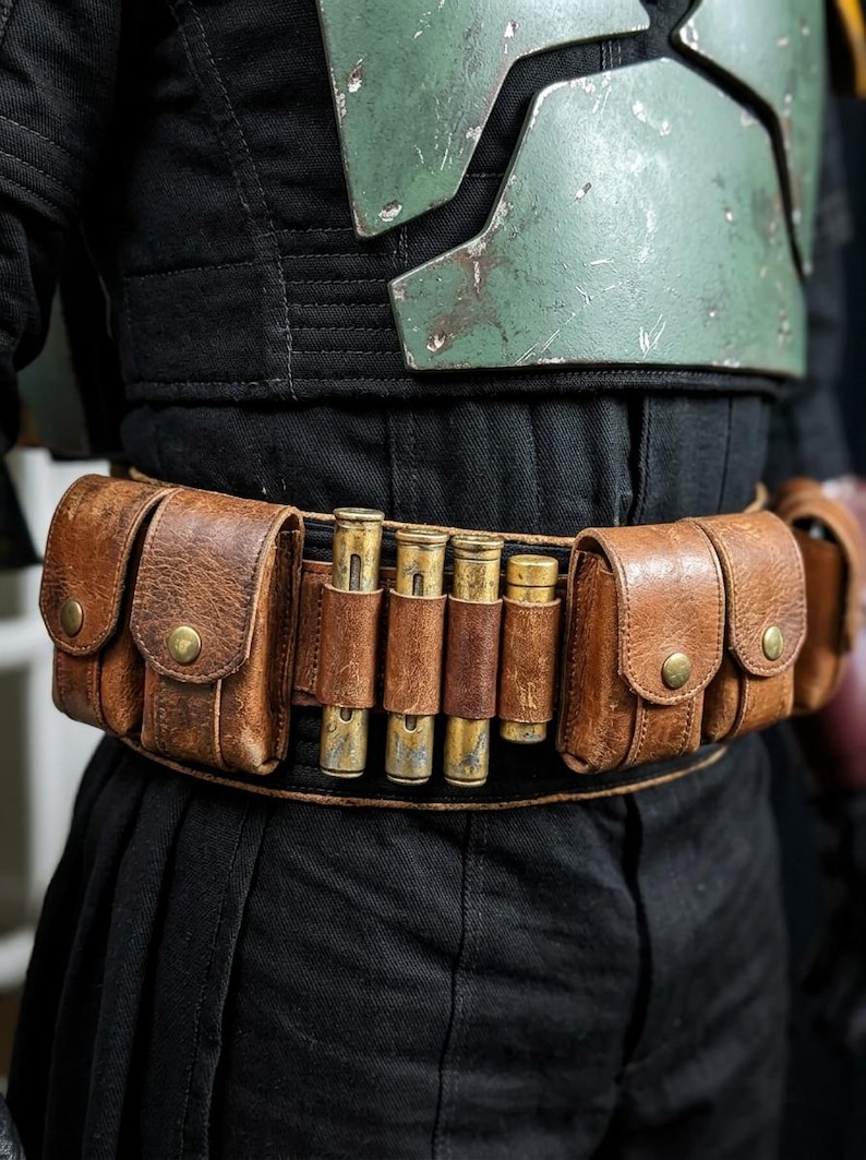 K&ouml;nnte beinhalten: Ein brauner Leder-Utility-G&uuml;rtel mit mehreren Taschen und messingfarbenen Nieten. Der G&uuml;rtel verf&uuml;gt &uuml;ber mehrere zylindrische Metallobjekte, die von Lederriemen gehalten werden. Der G&uuml;rtel wird &uuml;ber schwarzer Kleidung getragen, wobei eine gr&uuml;ne und silberne Brustplatte sichtbar ist.