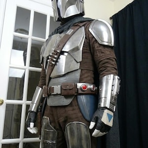 May include: A detailed costume resembling a Mandalorian warrior. The outfit features a silver helmet, chest armor, and gauntlets. Brown and gray fabrics make up the under-suit and pants. A utility belt and cape complete the look.