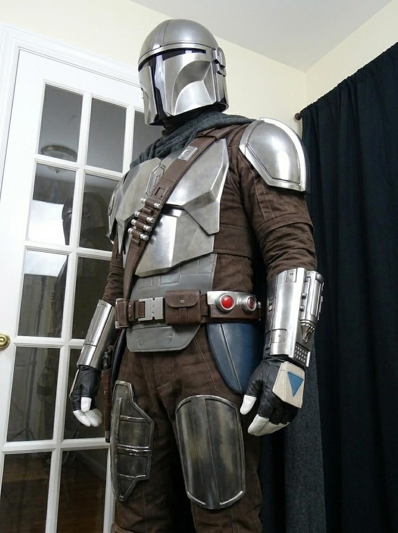 May include: A detailed costume resembling a Mandalorian warrior. The outfit features a silver helmet, chest armor, and gauntlets. Brown and gray fabrics make up the under-suit and pants. A utility belt and cape complete the look.