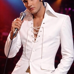 May include: A man in a white suit with a lace-up front, holding a microphone. The suit has a wide collar and is paired with white pants. A gold chain is visible. The background is blurred with stage lighting.