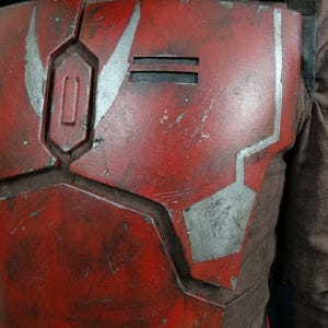 Puede incluir: Primer plano de una pieza de armadura de pecho roja y plateada. La armadura tiene un aspecto desgastado con detalles plateados y formas geom&eacute;tricas. La pieza del pecho es parte de un disfraz o cosplay.