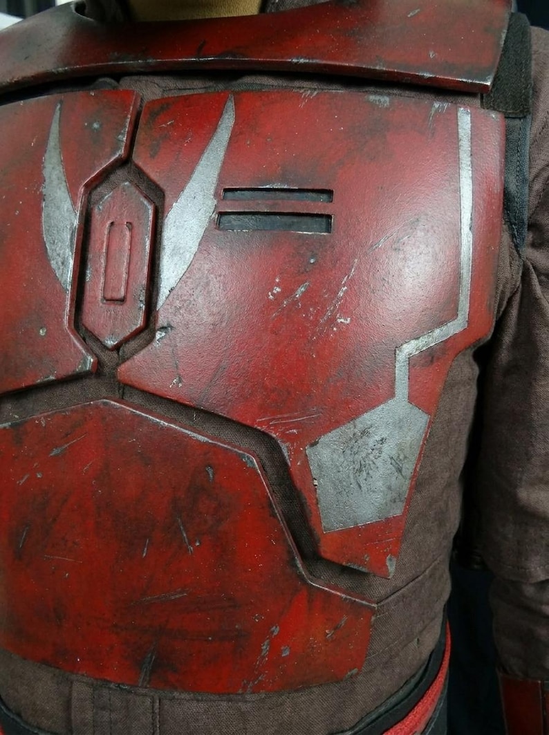 Puede incluir: Primer plano de una pieza de armadura de pecho roja y plateada. La armadura tiene un aspecto desgastado con detalles plateados y formas geom&eacute;tricas. La pieza del pecho es parte de un disfraz o cosplay.