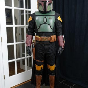 K&ouml;nnte beinhalten: Ganzk&ouml;rperkost&uuml;m, das Boba Fett aus Star Wars &auml;hnelt. Das Kost&uuml;m hat einen gr&uuml;nen Helm, eine Brustpanzerung und einen schwarzen Overall mit braunen Stiefeln. Details sind ein G&uuml;rtel mit Taschen, Panzerhandschuhe und Kniesch&uuml;tzer.