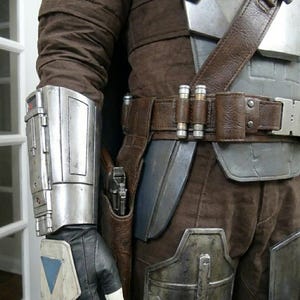 May include: A detailed view of a brown and silver costume, featuring a leather belt with ammunition holders and a holster. The costume includes a silver arm guard, brown pants, and a black glove with a white and blue accent. The overall theme is a futuristic or sci-fi design.