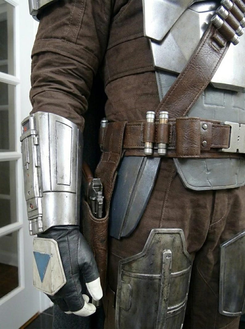 May include: A detailed view of a brown and silver costume, featuring a leather belt with ammunition holders and a holster. The costume includes a silver arm guard, brown pants, and a black glove with a white and blue accent. The overall theme is a futuristic or sci-fi design.