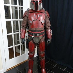 Puede incluir: Toma de cuerpo entero de una armadura mandaloriana roja y marr&oacute;n. El casco tiene una placa frontal plateada y cuernos. La armadura incluye petos, hombreras y armadura para las piernas. Se ve un cintur&oacute;n negro con una funda y una pistola bl&aacute;ster.