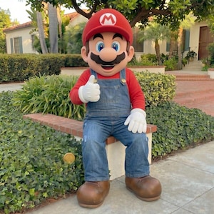 Pu&ograve; includere: Una persona in costume da Mario, indossa un cappello rosso con il logo "M" bianco, una maglietta rossa a maniche lunghe, una salopette di jeans blu e scarpe marroni. Il personaggio fa il gesto del pollice in su con guanti bianchi. Lo sfondo include vegetazione e un edificio.