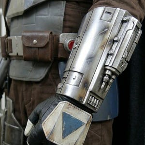 May include: Close-up of a silver and weathered metal gauntlet with a black glove. The gauntlet has a blue triangle design on the wrist and is part of a costume. A brown leather belt with a pouch is also visible.