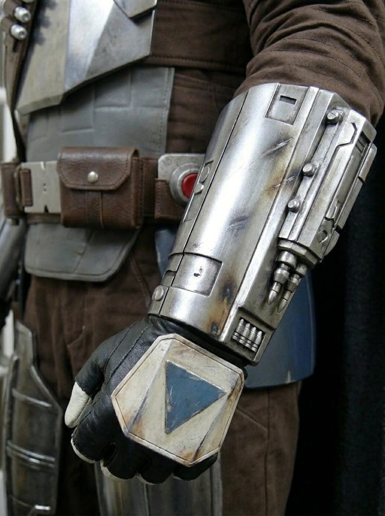 May include: Close-up of a silver and weathered metal gauntlet with a black glove. The gauntlet has a blue triangle design on the wrist and is part of a costume. A brown leather belt with a pouch is also visible.