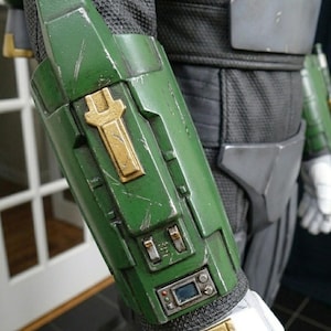 K&ouml;nnte beinhalten: Nahaufnahme eines gr&uuml;n-goldenen Armschutzes mit abgenutztem Aussehen. Der Schutz hat ein goldenes Emblem und kleine elektronische Bauteile. Der Armschutz ist Teil eines Kost&uuml;ms oder einer R&uuml;stung, mit einem schwarzen und grauen Stoff&auml;rmel sichtbar.
