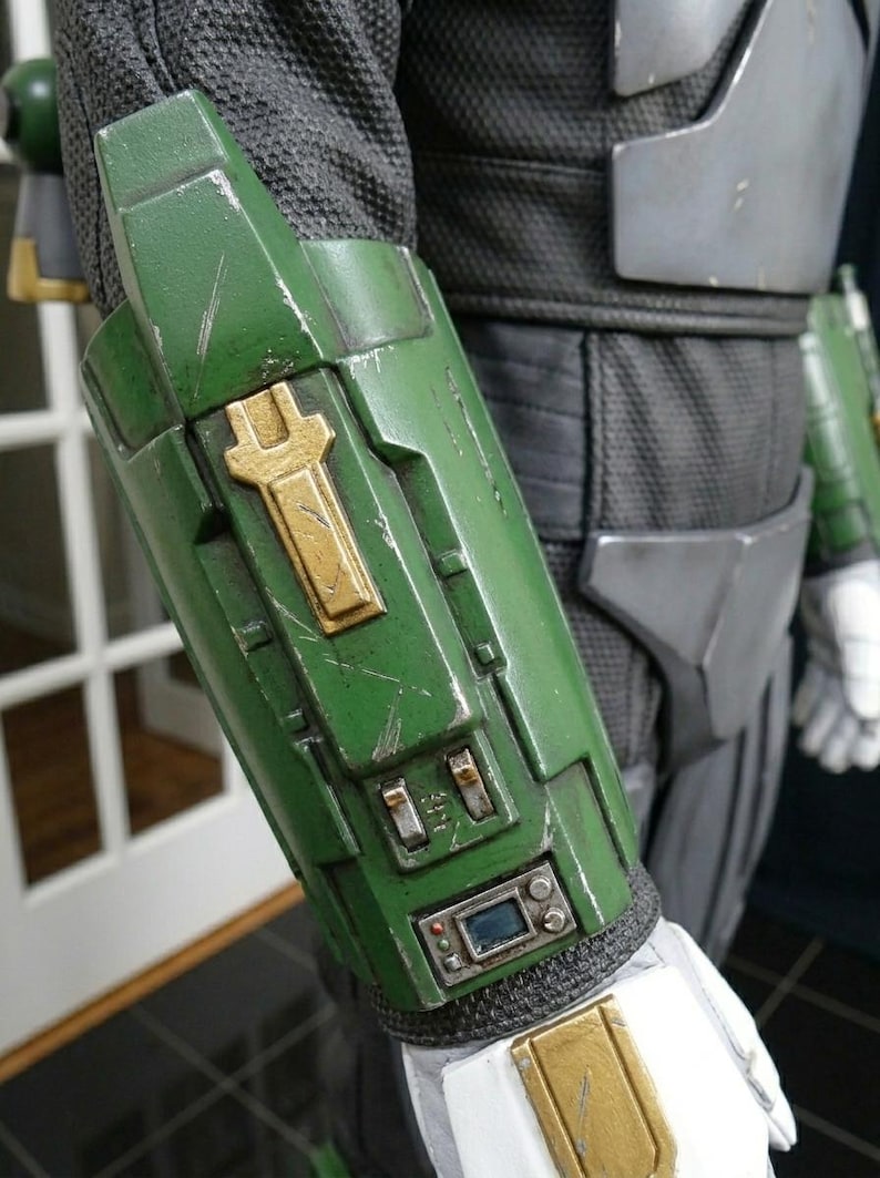 K&ouml;nnte beinhalten: Nahaufnahme eines gr&uuml;n-goldenen Armschutzes mit abgenutztem Aussehen. Der Schutz hat ein goldenes Emblem und kleine elektronische Bauteile. Der Armschutz ist Teil eines Kost&uuml;ms oder einer R&uuml;stung, mit einem schwarzen und grauen Stoff&auml;rmel sichtbar.