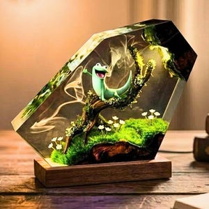 May include: A decorative lamp featuring a green lizard figurine inside a clear, geometric resin block. The resin encases a miniature landscape with moss, flowers, and a tree branch, set on a wooden base. The lamp is illuminated, creating a warm glow.