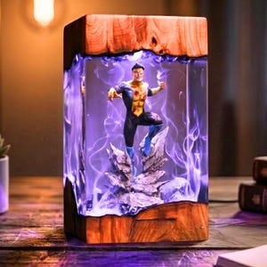 Puede incluir: Lámpara rectangular de madera y resina con una figura de superhéroe. La lámpara tiene un marco de madera con un centro de resina transparente, que muestra una figura de superhéroe sobre una formación rocosa, iluminada con luz púrpura.