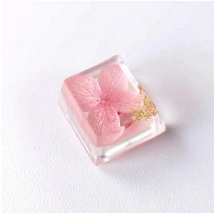 May include: A clear, rectangular resin piece encasing a delicate pink flower with gold flakes. The floral element is centered within the transparent block, creating a unique decorative item.