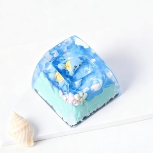 Può includere: Un cubo di resina trasparente con una scena oceanica in miniatura con un delfino blu e nuvole bianche. La base è azzurra con dettagli bianchi e rosa. Una conchiglia è posta di lato.