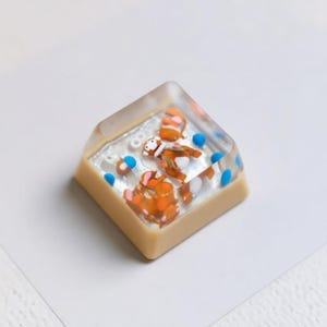 Pode incluir: Uma tecla personalizada com um design de resina transparente. Apresenta personagens de desenhos animados laranja e brancos e detalhes azuis. A tecla tem uma base bege.