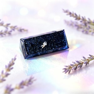 May include: A rectangular resin piece with a dark blue, glittery interior, featuring a tiny silver astronaut figure. The edges are a darker blue. Lavender sprigs are in the background, with a soft, iridescent light.