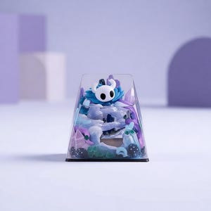 Könnte beinhalten: Ein klarer, trapezförmiger Behälter gefüllt mit bunten Miniaturfiguren. Die Figuren umfassen eine weiße Figur mit großen schwarzen Augen und eine blaue blütenartige Struktur. Weitere Elemente sind Schattierungen von Lila, Blau und Weiß.