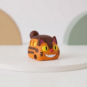 Puede incluir: Figura con forma de gato naranja y marrón con grandes ojos amarillos y boca sonriente. La figura tiene forma redondeada con rayas marrones y parte superior marrón. El gato tiene una cola pequeña y redonda marrón y orejas internas rosas.