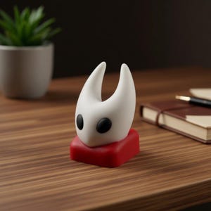 Könnte beinhalten: Eine weiße Figur mit schwarzen Augen und zwei nach oben gebogenen Hörnern, die auf einem roten rechteckigen Sockel steht. Die Figur steht auf einer Holzoberfläche, im Hintergrund ein kleiner Blumentopf und ein Notizbuch.