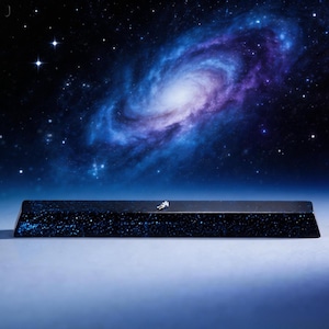 May include: A dark, rectangular object with a starry pattern, possibly a keyboard or desk accessory, featuring a miniature astronaut figure. The background showcases a vibrant galaxy with swirling blues, purples, and white stars.