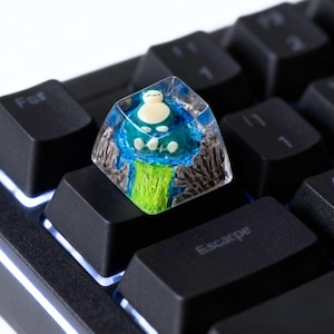 May include: A custom keycap featuring a Snorlax design. The keycap is a clear, square shape with a detailed, colorful depiction of Snorlax on a blue and green landscape. The keycap is installed on a black mechanical keyboard.