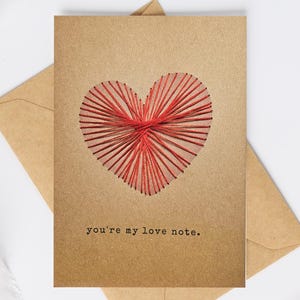 May include: A kraft paper greeting card featuring a stitched red heart design. The card has the text "you're my love note." An envelope is visible behind the card, suggesting a romantic theme.