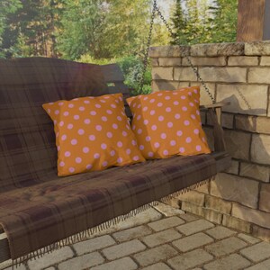Könnte beinhalten: Zwei orangefarbene Kissen mit rosa Punkten liegen auf einer Holzschaukel. Die Schaukel hat eine braune karierte Decke. Der Hintergrund umfasst eine Backsteinmauer, Bäume und eine gepflasterte Terrasse, die eine gemütliche Umgebung im Freien schaffen.