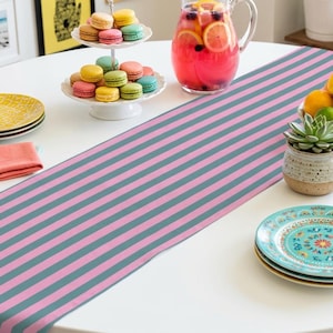 May include: A table setting featuring a pink and gray striped table runner. A two-tiered serving dish holds colorful macarons. A pitcher of pink lemonade with fruit slices sits nearby, along with a small succulent and decorative plates.