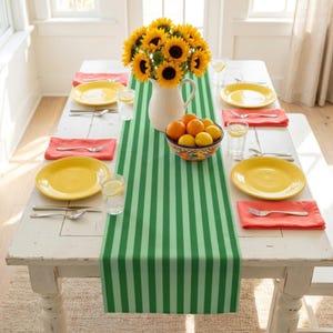 May include: A dining table set for a meal with a green and white striped table runner. Yellow plates, orange napkins, and silverware are set for each place. A vase of sunflowers and a bowl of citrus fruits complete the table setting.