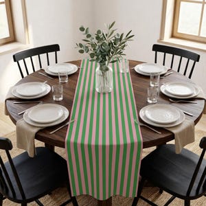 May include: A round wooden dining table set for six, featuring a green and pink striped table runner. Each place setting includes a white plate, silverware, and a clear glass. A vase of greenery sits in the center.