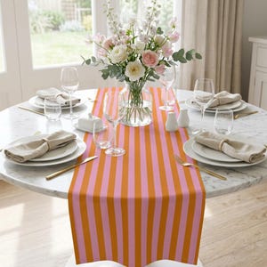 Puede incluir: Una mesa redonda de mármol preparada para una comida, con un camino de mesa a rayas naranja y rosa vibrante. La mesa está puesta con platos blancos, cubiertos dorados y copas de vino. Un jarrón de flores añade un toque de elegancia.