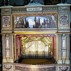 Pode incluir: Um palco de teatro em miniatura detalhado com entalhes ornamentados e detalhes pintados. O palco apresenta um cenário pintado de uma paisagem, com cortinas drapeadas e colunas decorativas. A parte superior do palco tem a inscrição "ANNO MCMXVIII".