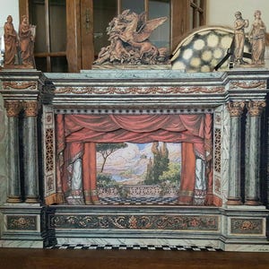 May include: A detailed diorama of a classical theater stage. The stage features red curtains, painted backdrop of a landscape, and decorative columns. Statues and a Pegasus sculpture adorn the top. The diorama is set on a wooden surface.
