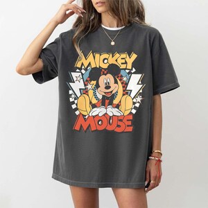 May include: Dark gray t-shirt featuring a colorful graphic of Mickey Mouse. The design includes the words "Mickey Mouse" in yellow and red, with lightning bolts and stars. The shirt has short sleeves and a crew neck.