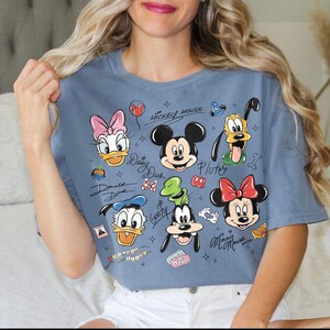 May include: A blue t-shirt featuring cartoon illustrations of Mickey Mouse, Minnie Mouse, Donald Duck, Daisy Duck, Goofy, and Pluto. The shirt has the characters' names and the words "Mickey Mouse" and "Magic" printed on it.