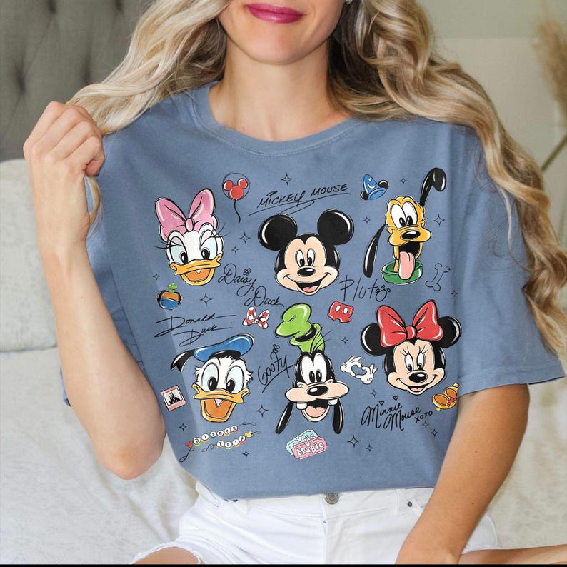 May include: A blue t-shirt featuring cartoon illustrations of Mickey Mouse, Minnie Mouse, Donald Duck, Daisy Duck, Goofy, and Pluto. The shirt has the characters' names and the words "Mickey Mouse" and "Magic" printed on it.
