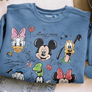 May include: A light blue sweatshirt featuring Mickey Mouse and friends. The design includes illustrations of Mickey, Minnie, Donald, Daisy, Goofy, and Pluto. The words "Mickey Mouse" and character names are written in a script font.