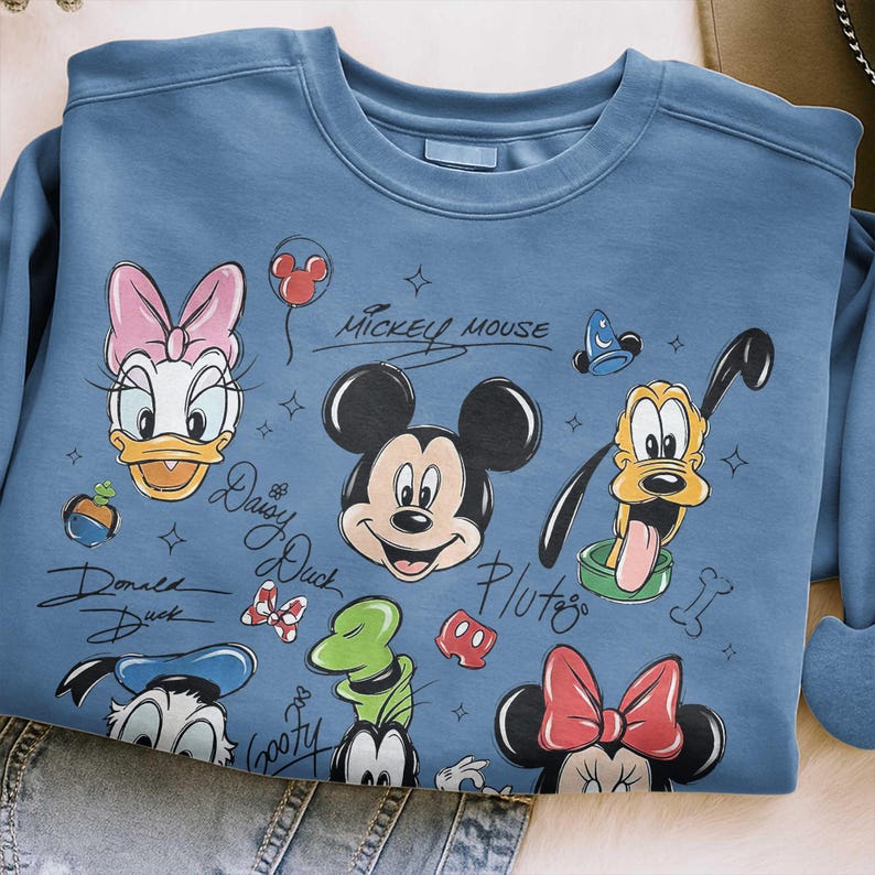 May include: A light blue sweatshirt featuring Mickey Mouse and friends. The design includes illustrations of Mickey, Minnie, Donald, Daisy, Goofy, and Pluto. The words "Mickey Mouse" and character names are written in a script font.